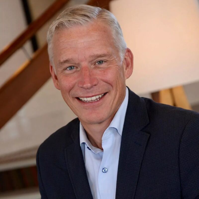 The image shows a middle-aged man with short, gray hair and a friendly smile. He is wearing a light blue shirt under a dark blue blazer. The background is slightly blurred, suggesting an indoor setting, possibly an office or a home. The man appears to be looking directly at the camera.